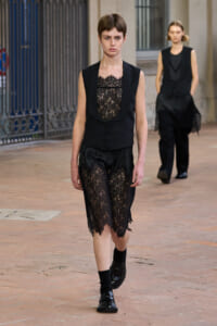 Model walking a runway in a black lace dress with a satin vest, wearing black shoes on a brick courtyard, background model blurred out