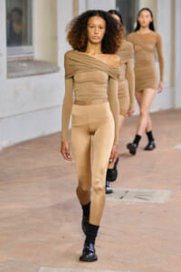Model walks a fashion runway in beige off-shoulder top and matching leggings; other models in similar outfits follow behind.