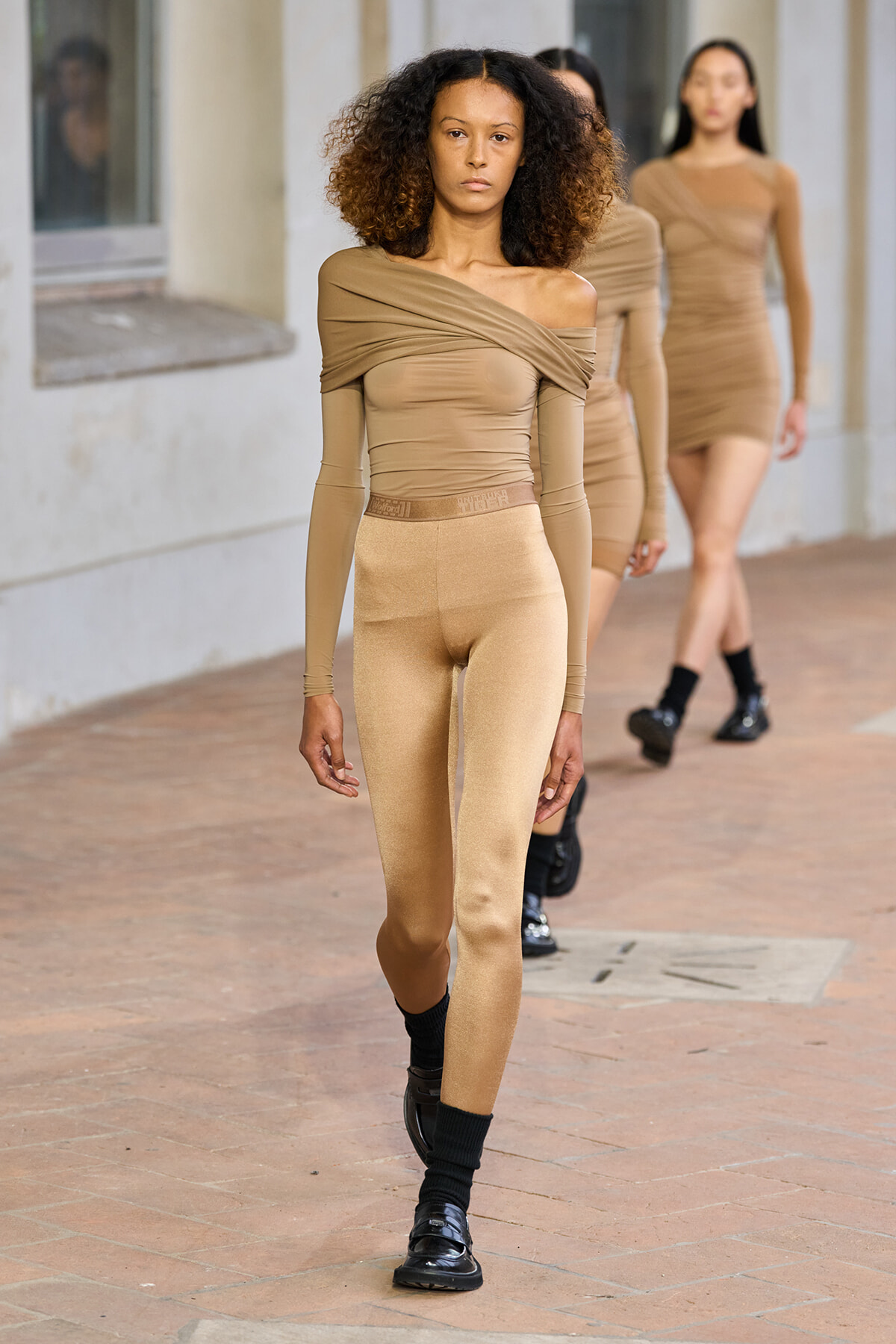 Model walks a fashion runway in beige off-shoulder top and matching leggings; other models in similar outfits follow behind.