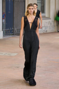 Model walking a street-style runway in a black lace-trimmed jumpsuit with a plunging neckline and wide legs, wearing fuzzy black slippers.