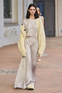 Model on a fashion runway wearing a pale beige satin jumpsuit with an oversized cream knitted cardigan draped over her shoulders.