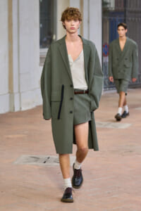 Male model walks down a street in an oversized olive-green coat, beige henley, brown belt, and black lace-up shoes.