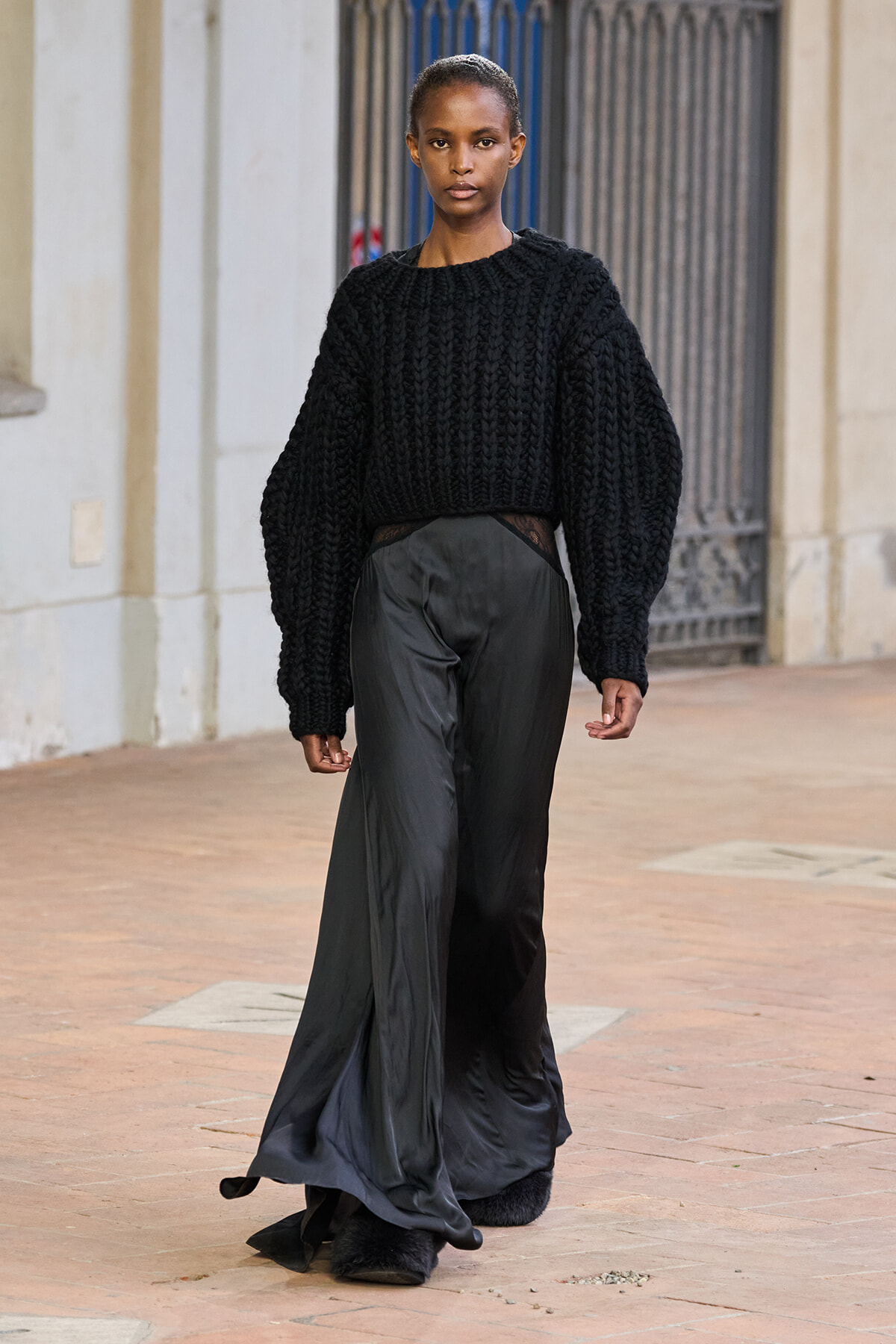 Model walking outdoors in a bulky black knit sweater and wide, satin-black pants, looking straight ahead on a brick-paved street.