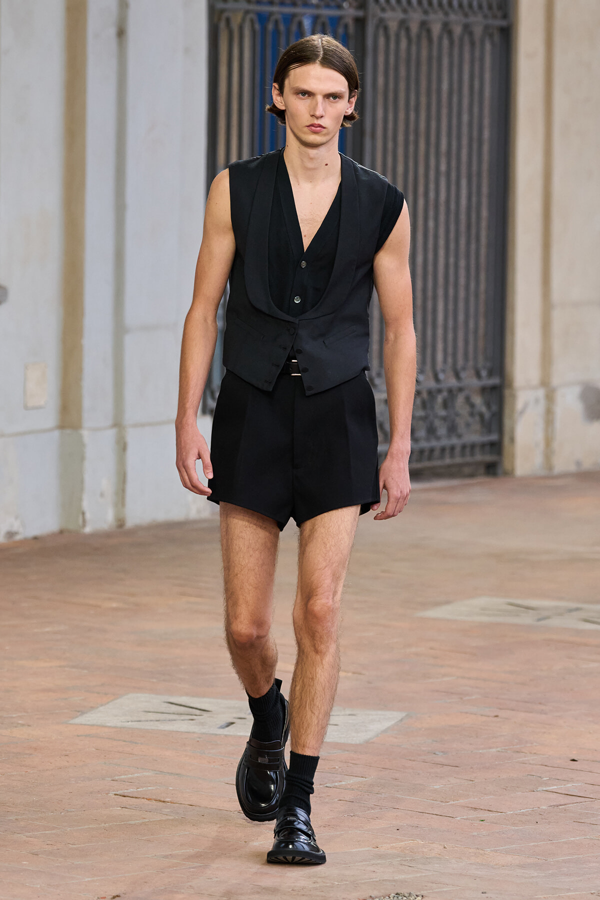Male runway model walking in a black sleeveless vest, matching vest and shorts, with black socks and shiny loafers on a brick walkway.