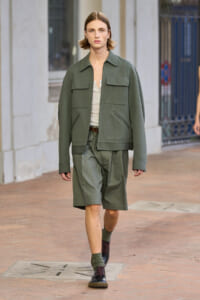 Male model walking on a city street in an olive green utility jacket and matching shorts with a beige top