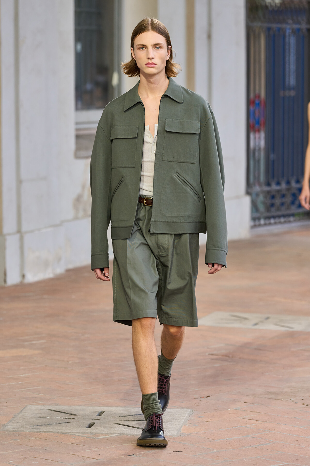Male model walking on a city street in an olive green utility jacket and matching shorts with a beige top