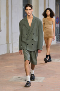Male model walks the runway in an oversized olive-green double-breasted blazer with matching shorts; a beige dress-clad model follows behind.