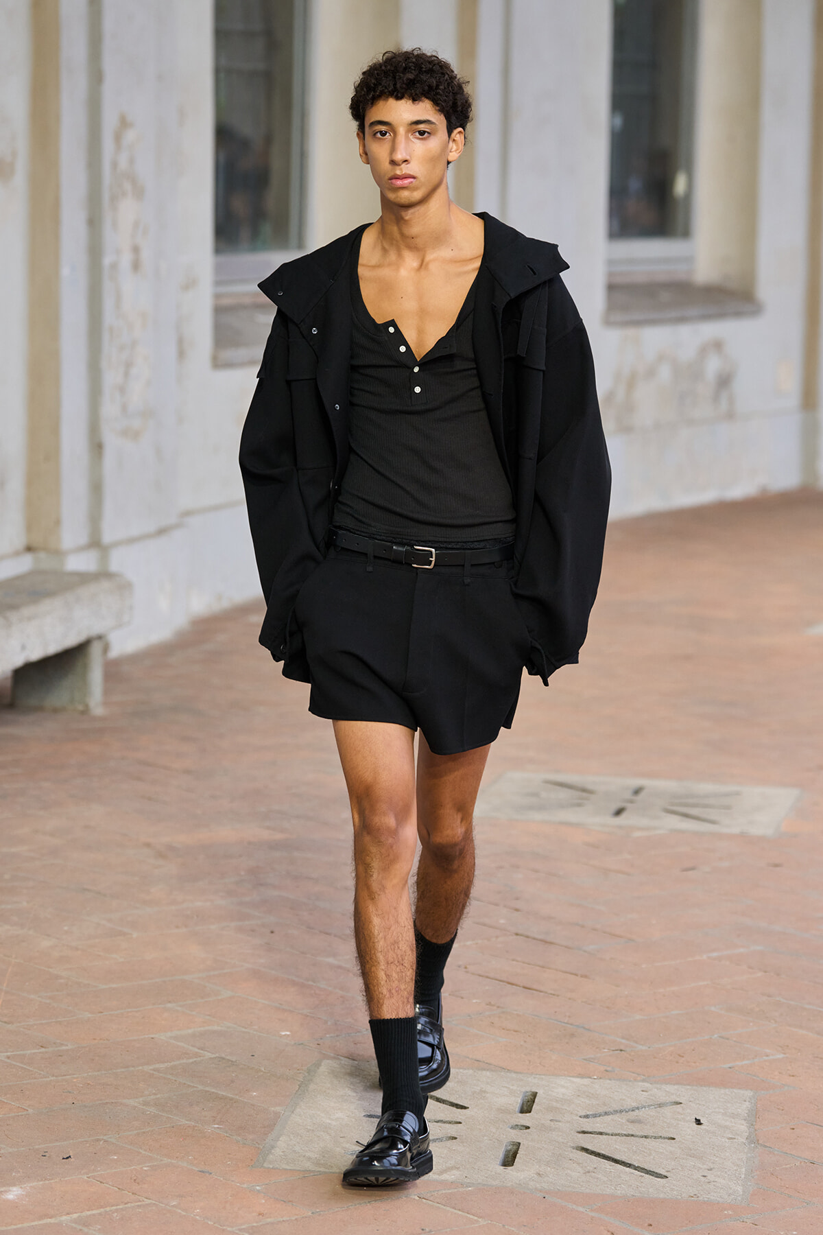 Young man in an all-black outfit walks along a brick sidewalk near a beige stone building, wearing a loose jacket, black henley, belt, and black loafers with socks.