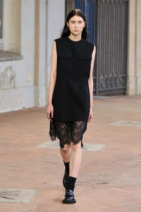 Female model wearing a sleeveless black dress with two front pockets and a lace-hemline, walking on a brick city sidewalk.