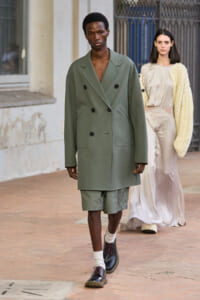 Male model in an oversized sage-green double-breasted coat with matching shorts, burgundy shoes, and cream socks walking a runway.