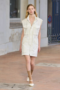 Female model walking on a city street in a sleeveless white utility dress with large front pockets and a front zipper.