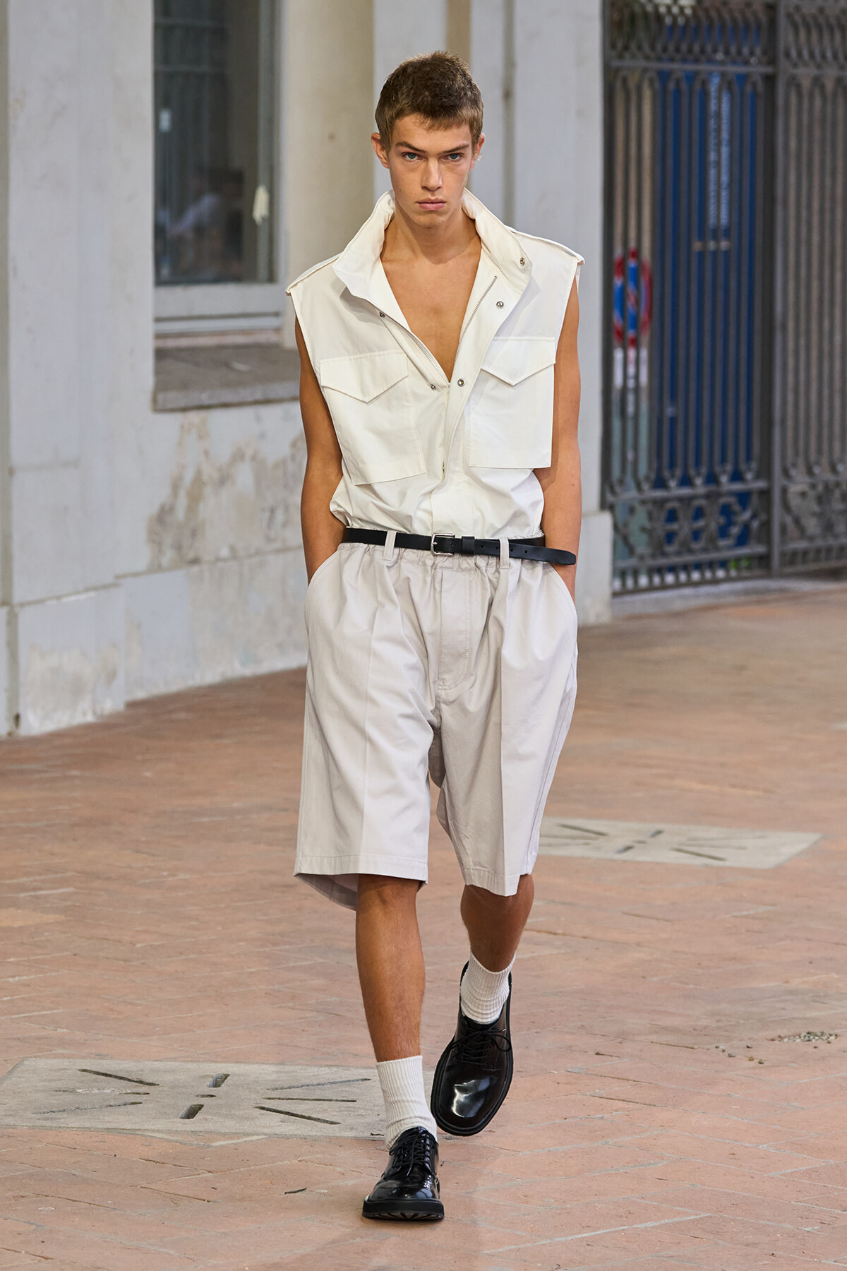 Male fashion model walking outdoors in a white sleeveless shirt and knee-length shorts with a black belt.