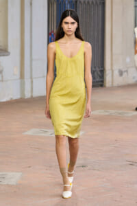 Model walking outdoors in a yellow sleeveless slip dress on a brick sidewalk with a gated building in the background