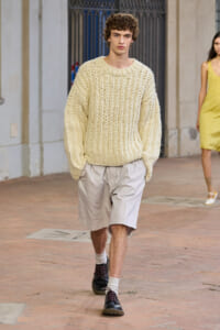 Male model walks a runway in an oversized cream cable-knit sweater with light beige shorts, white socks, and dark lace-up shoes outdoors in a fashion show venue.