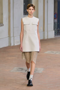 Model walking in a white sleeveless tunic dress with pocket flaps over a beige lace hem, black shoes, and ribbed socks in an outdoor set piece.