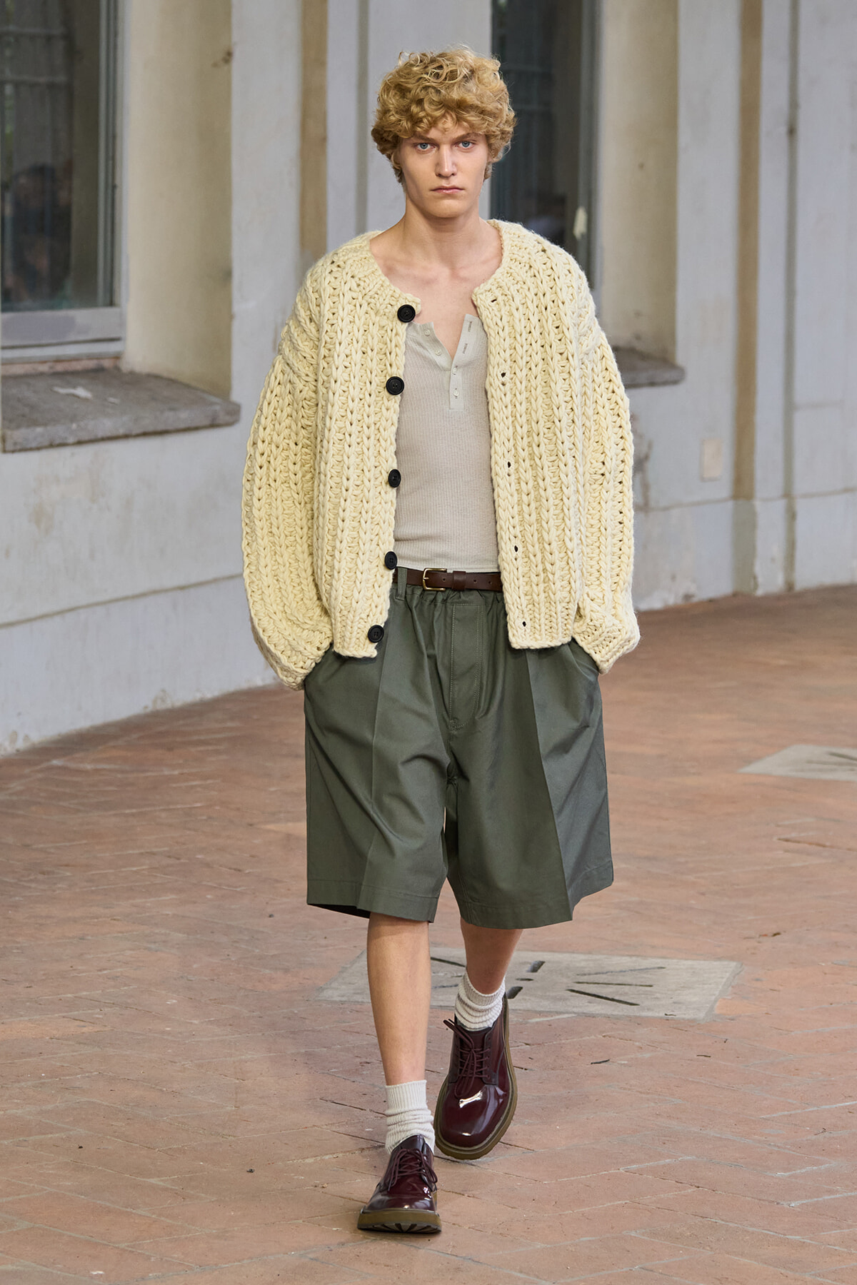 Male model walking outdoors in a chunky cream cardigan over a beige henley and olive shorts, burgundy shoes on a brick street.