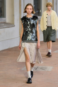 Model walks a fashion runway in a black, sequin-embellished sleeveless top and a pale satin skirt with lace trim; another model in a cream cardigan walks behind.