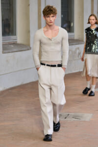 Male model wearing a beige long-sleeve Henley with lace trim at the hem and cream wide-leg trousers with a black belt, black loafers, walking on a brick-paved outdoor runway.