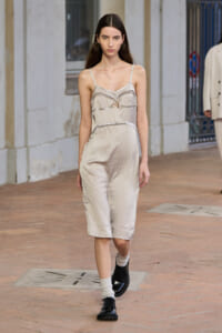 Model walking a runway in a beige satin slip dress with silver piping and a small bow at the chest, paired with white socks and black lace-up shoes.