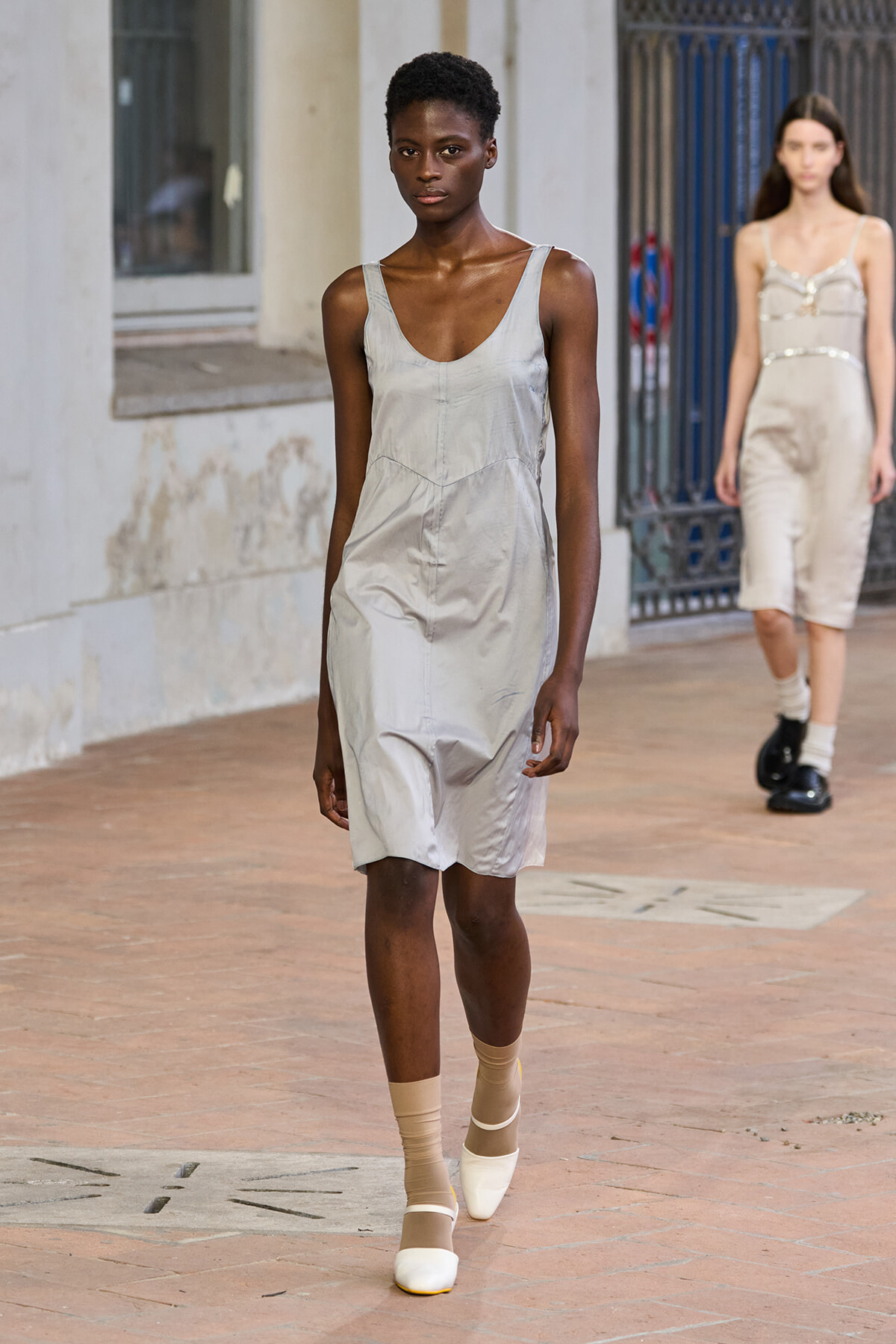 Model walking a runway in a light gray sleeveless dress, beige socks, and white shoes on a brick-paved catwalk with a companion in the background.