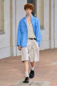 Young man with curly red hair walks on a brick sidewalk wearing a bright blue windbreaker, beige shirt, and knee-length khaki shorts with black shoes.