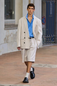 Male model in an oversized beige blazer and matching shorts, blue shirt, white socks, and black loafers walking on a city sidewalk.