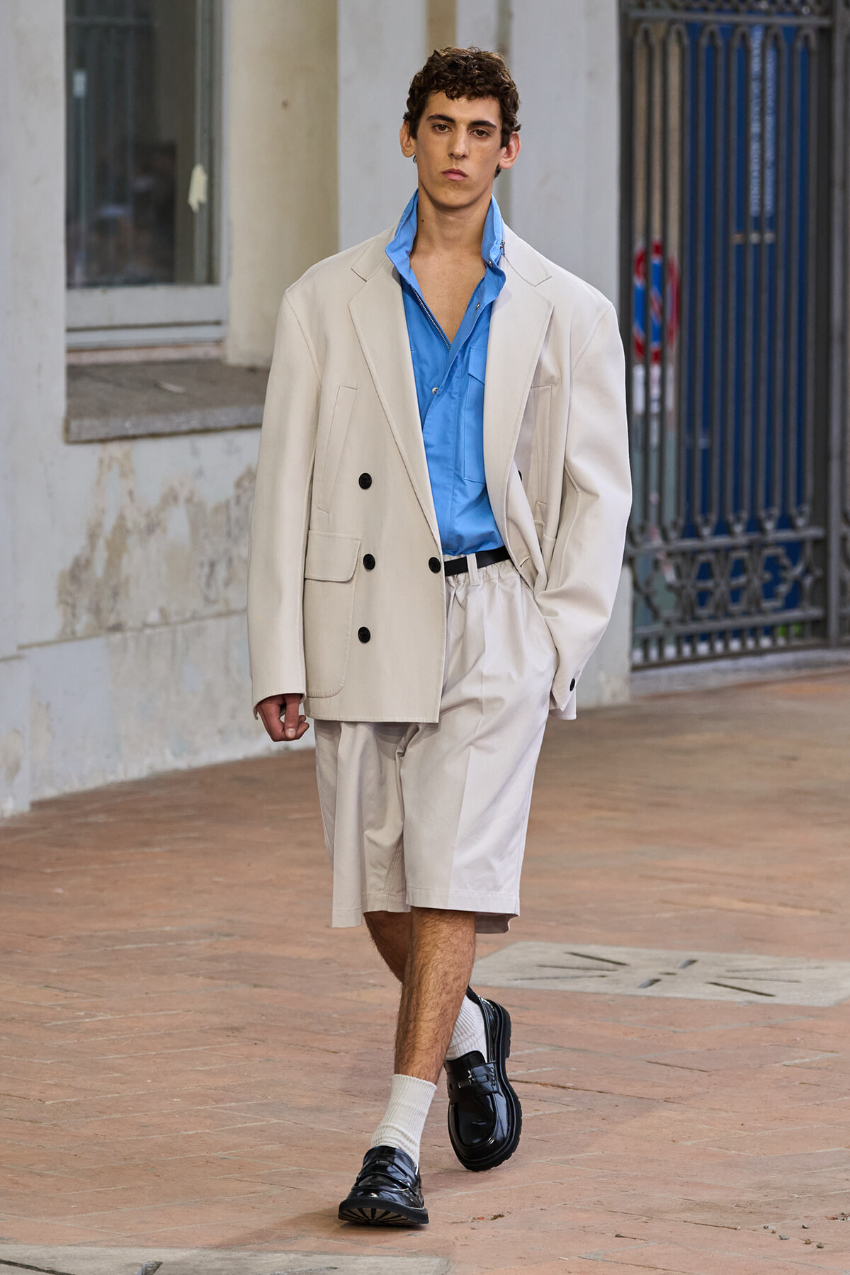 Male model in an oversized beige blazer and matching shorts, blue shirt, white socks, and black loafers walking on a city sidewalk.