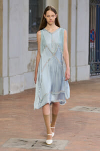 Female model walking on a brick street in a pale blue sleeveless dress with crystal bead trim and an uneven hem.