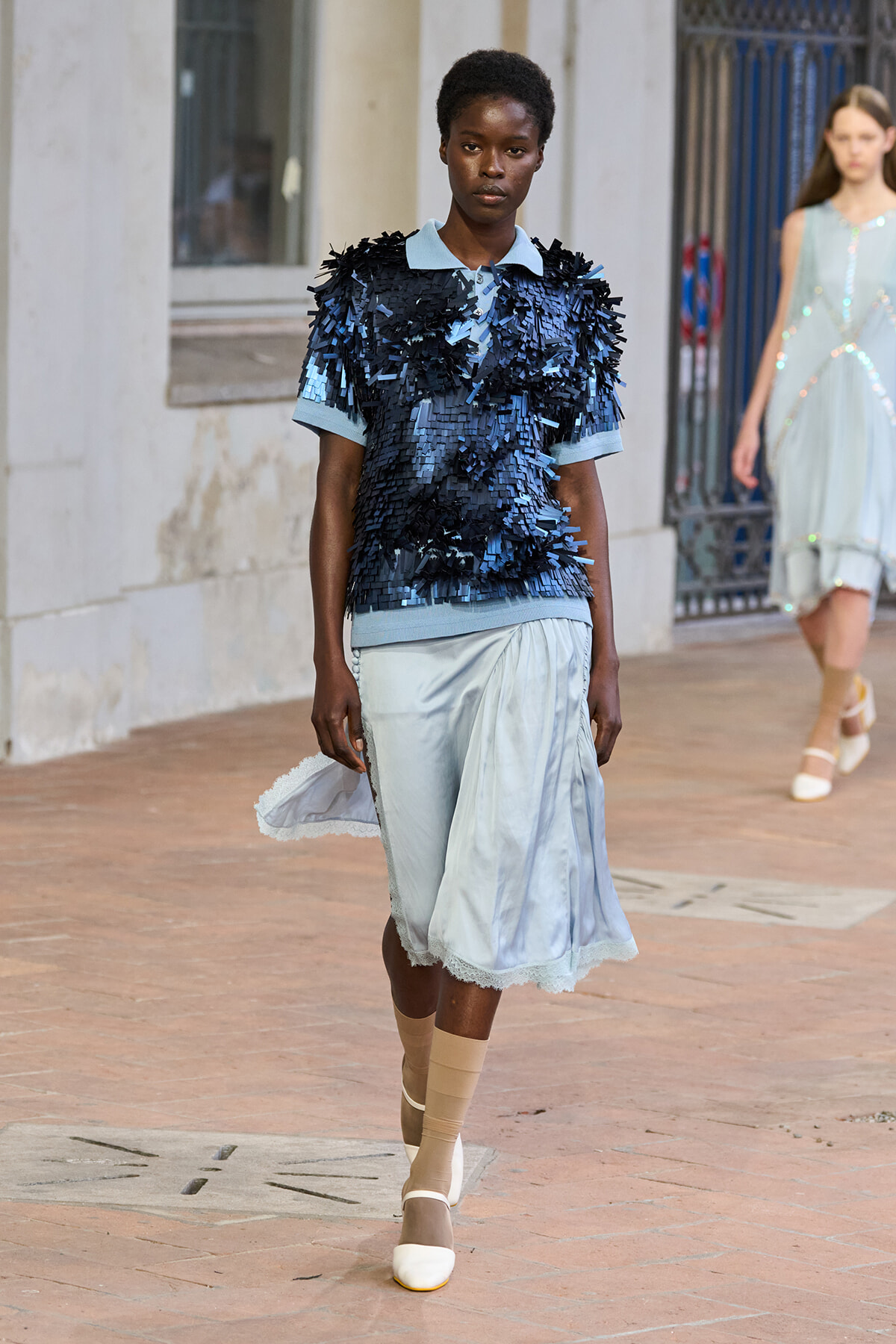 Model walks a runway wearing a navy fringed top over a light blue midi skirt with lace hem, and beige socks with white heels.