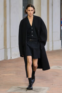 Model walking in an all-black, oversized outfit: long coat over a dark cardigan and black shorts on a city sidewalk.