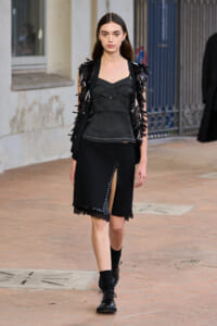 Model walking on a runway in an all-black outfit: a fringed vest over a structured top and a knee-length skirt with a front slit.