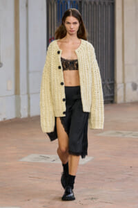 Model walking on a street in a chunky cream knit cardigan over a black lace bralette and a black satin skirt with lace trim, mid-thigh exposed on one side.