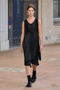 A female model walks down a city street wearing a sleeveless black dress with beaded geometric panels, black socks, and shiny loafers.