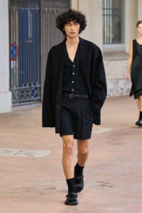 Male fashion model walking outdoors in an all-black outfit: blazer, cardigan-like outer layer, belt, shorts, and black sandals on a brick sidewalk.