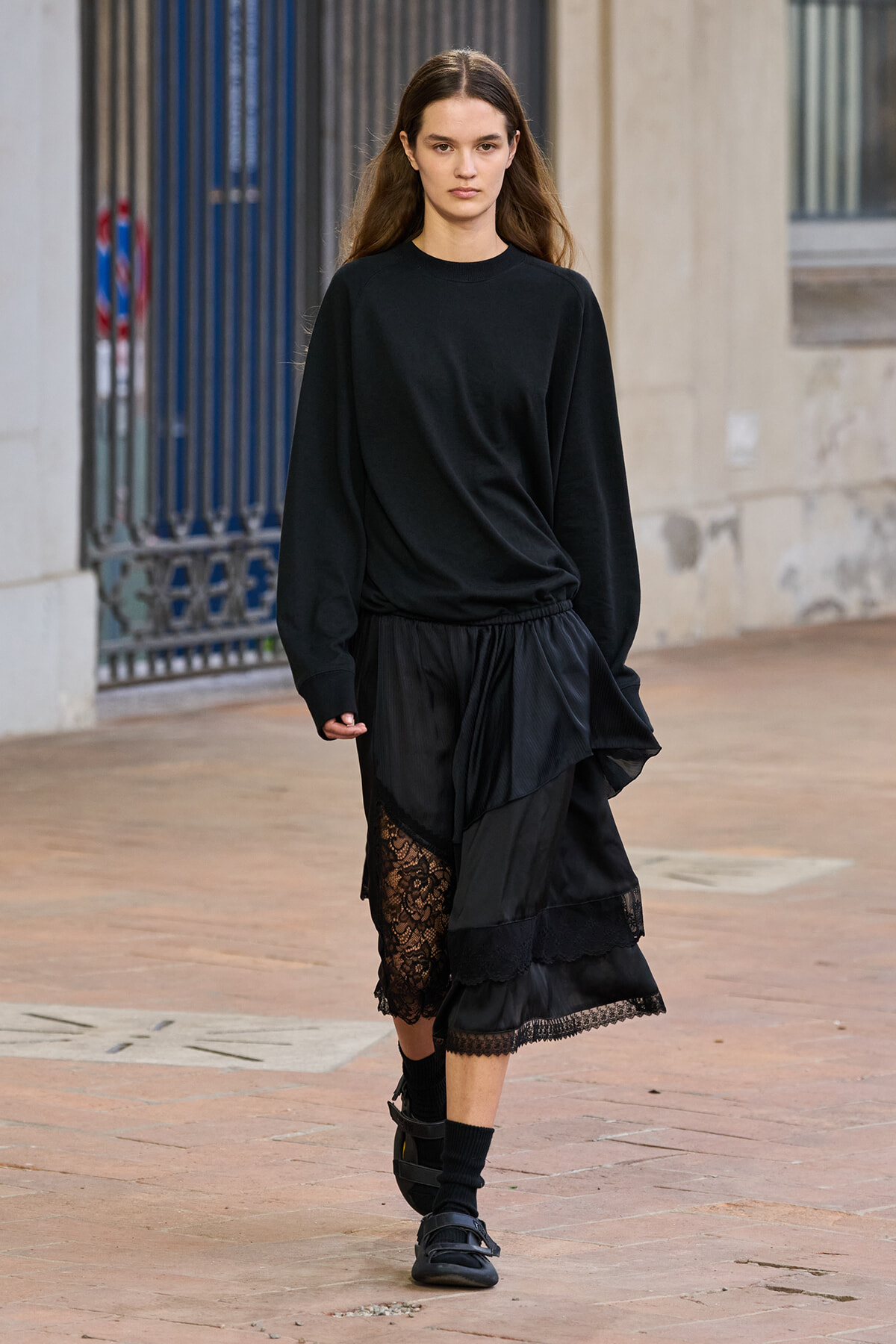 Young woman walking on a city sidewalk in an all-black outfit, layered skirt with lace hem visible at the knee depth behind her.