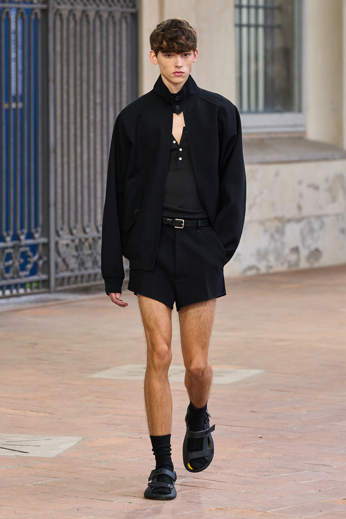 Male model walking on a brick sidewalk in an all-black outfit: short bomber/jacket, black polo-style top, belted black shorts, black socks, and strappy sandals.