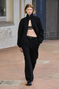 Model wearing an oversized black jacket, black loose trousers, and lace bralette, walking on a brick sidewalk with a serious expression.