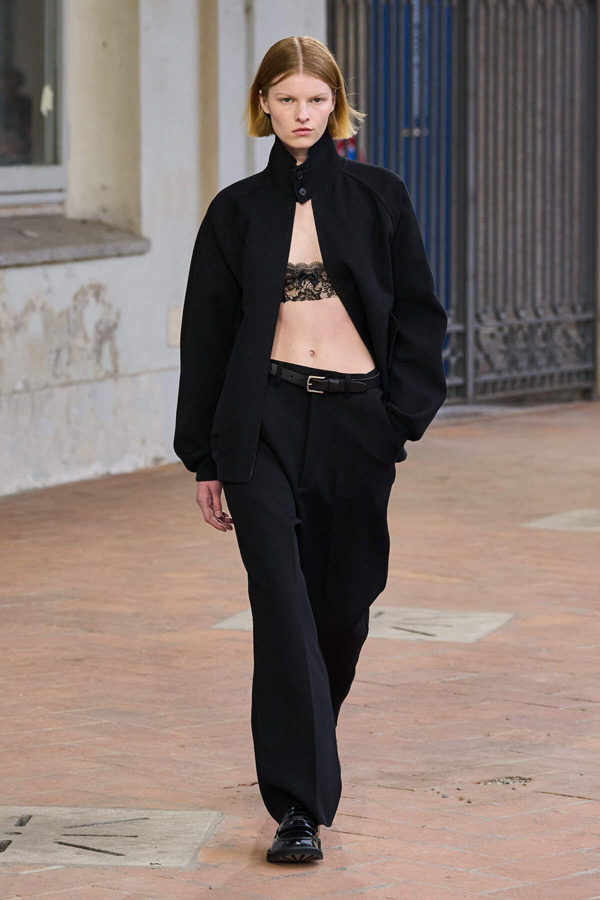 Model wearing an oversized black jacket, black loose trousers, and lace bralette, walking on a brick sidewalk with a serious expression.