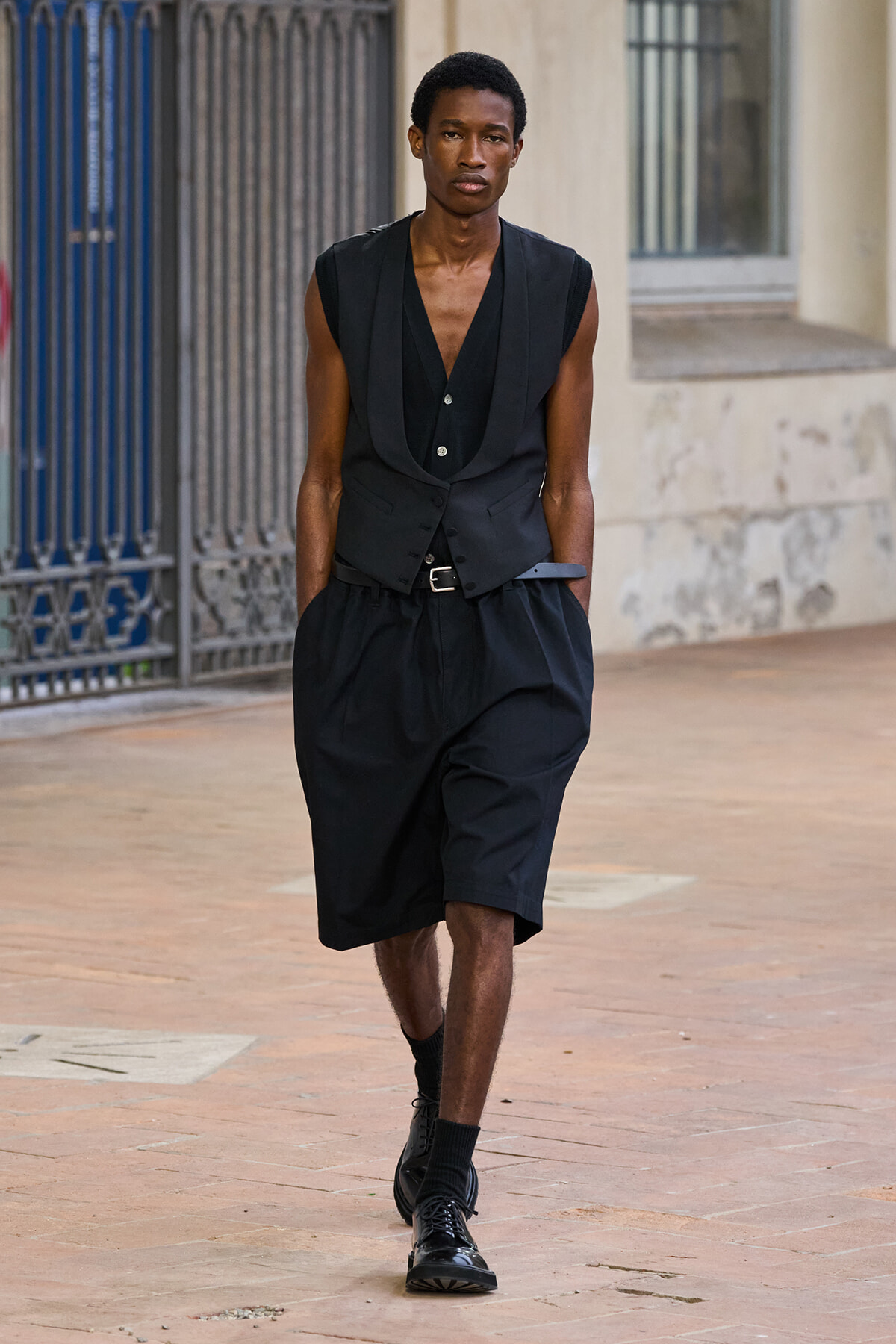 Slim man in an all-black outfit (sleeveless vest over a deep V-neck top, knee-length shorts, belt) walking on a city sidewalk.