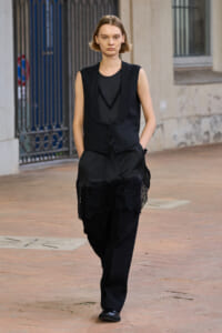 Model walking outdoors in a black monochrome outfit: sleeveless vest over a sheer top with lace details and lace-trimmed, loose black trousers. Hands in pockets on a brick-paved street.