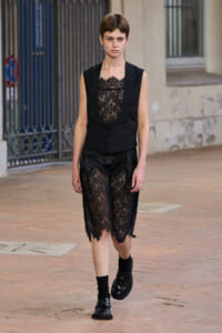 Model walking in a black lace outfit: sleeveless vest over sheer lace dress, outdoors on a brick-paved street.