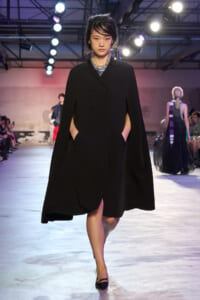 Model walks a fashion runway in a long black cape-style coat with hand pockets, under bright stage lights and a visible audience in the background.