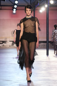 Model walking a runway in a sheer black outfit with a bejeweled collar, sheer top and flowing sheer pants, black heels.