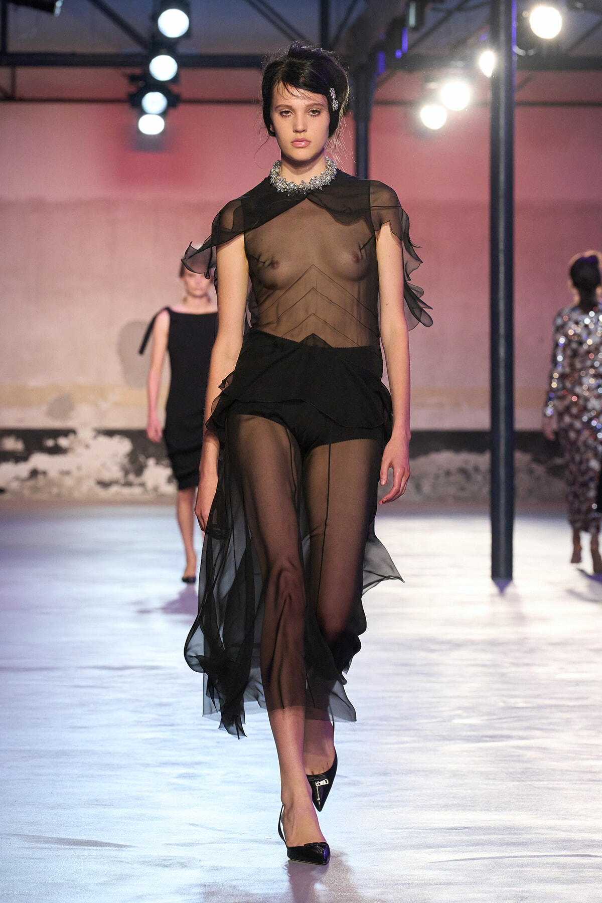Model walking a runway in a sheer black outfit with a bejeweled collar, sheer top and flowing sheer pants, black heels.