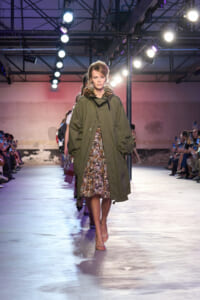 Model walks the runway wearing an olive green oversized parka with a fur-trimmed hood over a gold sequin dress, with bright lights and an audience on both sides.