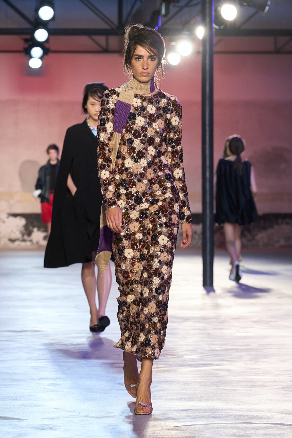 Model walking the runway in a full-length sequined floral dress with a beige and purple scarf, under bright stage lights.