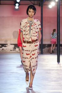 Model walks the runway in a cream cherry-print outfit with a striped top, brown belt, and long pearl necklaces, white flats.