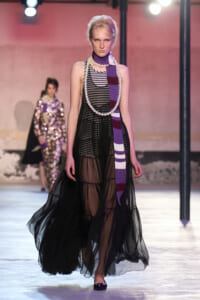 Model walks a fashion runway in a sheer black tiered gown, striped top detail, and a long purple-and-white scarf with a chunky pearl necklace.