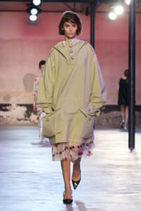 Model walking runway in oversized beige parka with a floral-hem dress peeking underneath, hands in pockets.