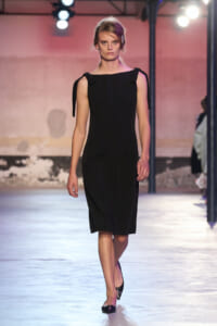 Model walks the runway in a knee-length black sheath dress with tied shoulder straps.